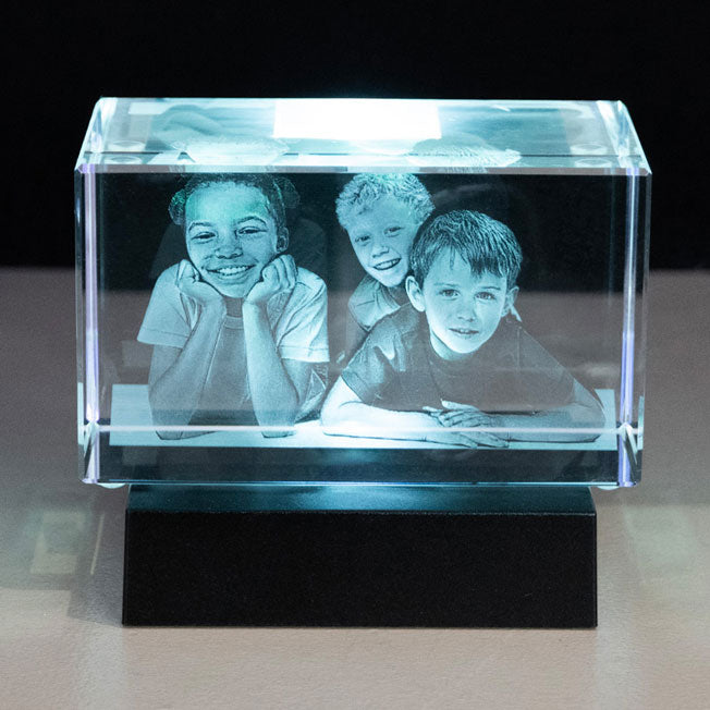 Bloc en verre gravé en 3D représentant trois enfants souriants, posé sur un socle lumineux noir qui fait ressortir les détails de la gravure.