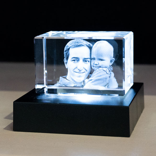 Bloc en verre gravé en 3D avec le portrait d’un père et de son bébé, posé sur un socle lumineux blanc, créant un rendu tendre et éclatant.