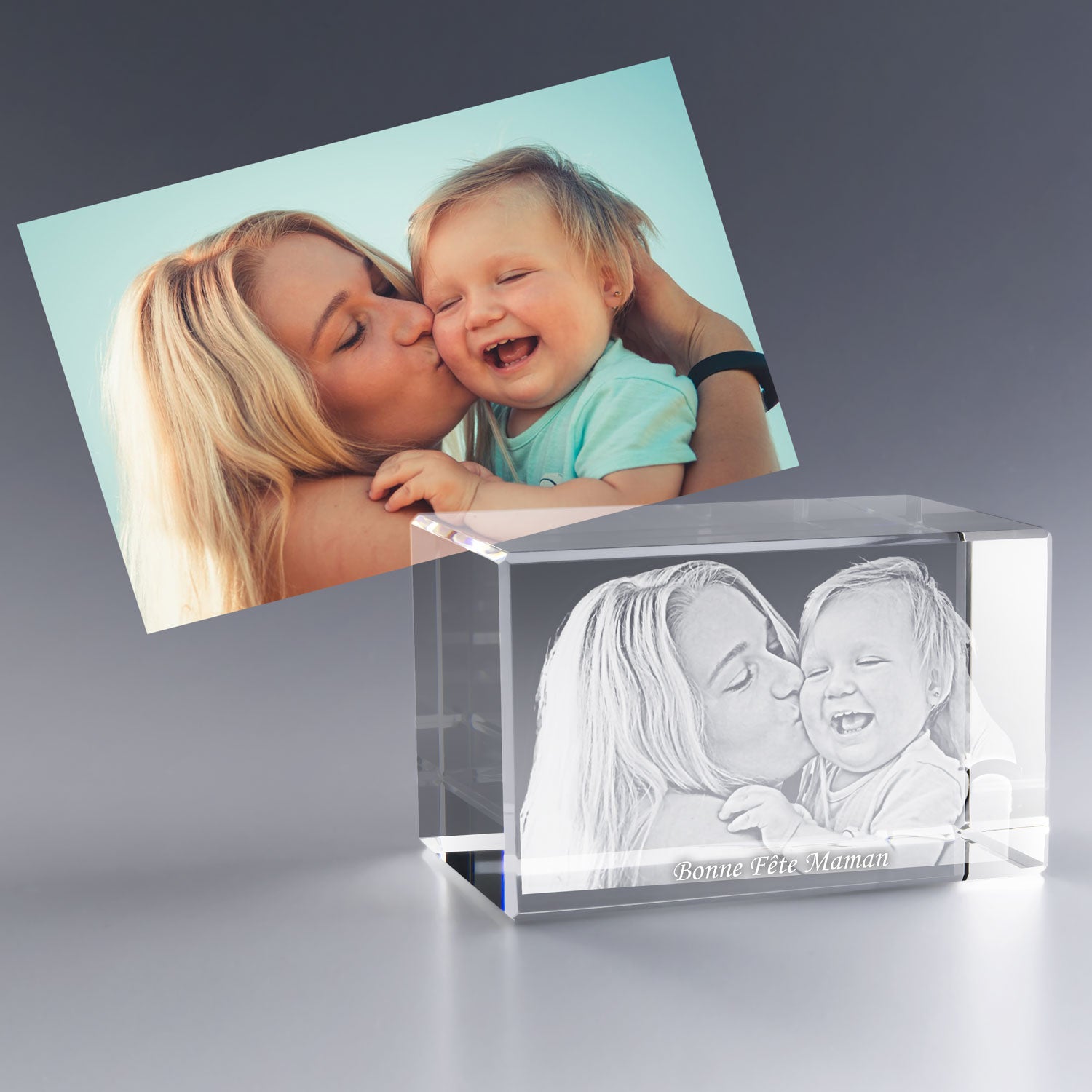 Bloc en verre gravé en 3D avec une mère embrassant tendrement son bébé, accompagné du texte “Bonne Fête Maman”, d’après la photo placée derrière.