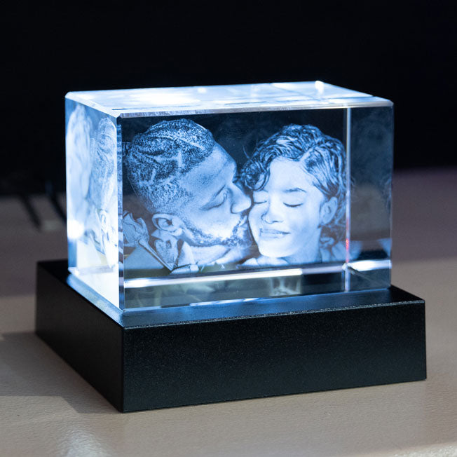 Bloc en verre gravé en 3D avec le portrait d’un couple complice, éclairé par un socle lumineux bleu qui révèle les détails de la gravure.