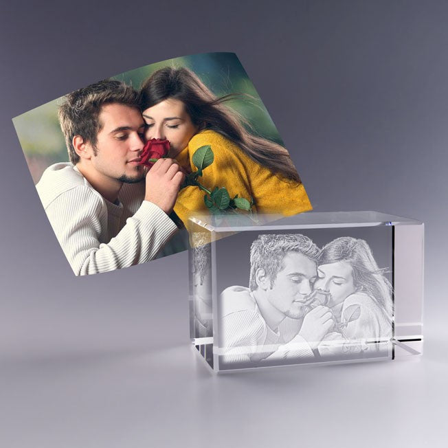 Bloc en verre gravé en 3D avec un couple tendre partageant une rose, d’après une photo couleur posée à l’arrière, ambiance douce et florale.