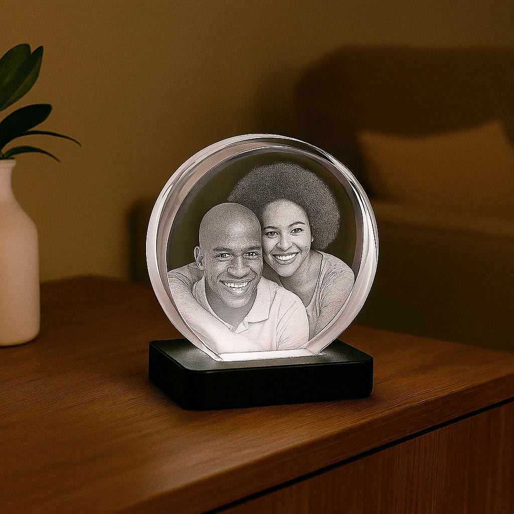 Bloc en verre rond gravé en 3D avec un portrait de couple, posé sur un socle lumineux blanc, sur une table de chevet en bois.