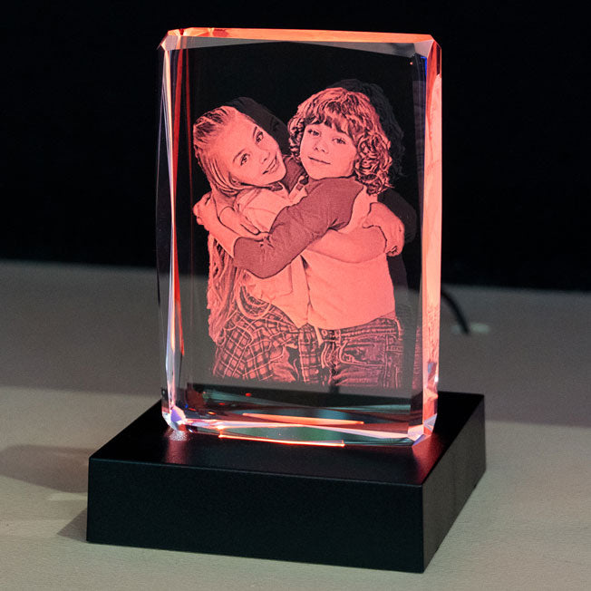 Bloc en verre vertical facetté rétroéclairé en rouge, avec la gravure 2D de deux enfants qui s’enlacent tendrement et sourient.