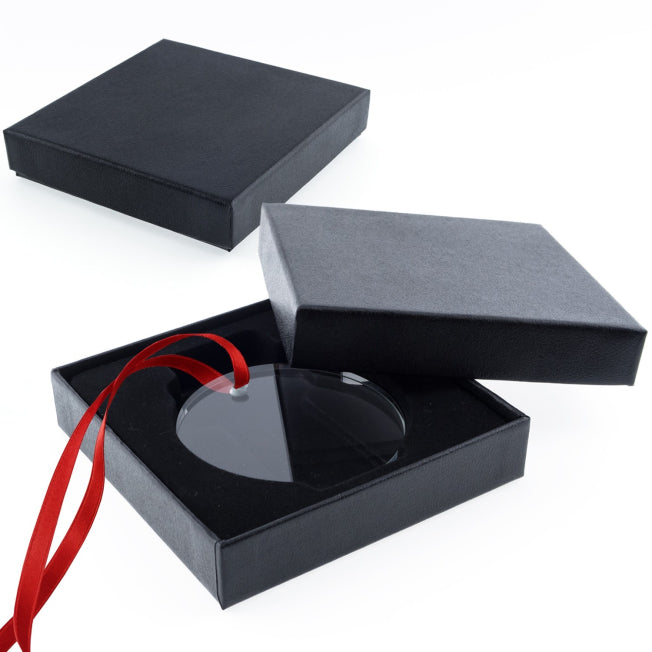 Boîte cadeau carrée noire ouverte, contenant un médaillon rond en verre avec un ruban rouge, sur fond blanc.
