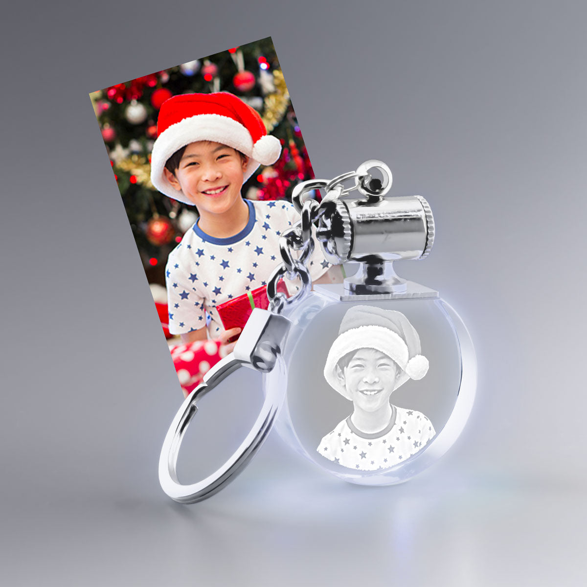 Porte-Clés en Verre Lumineux Rond avec photo gravée 2D – Cadeau de Noël personnalisé – Un enfant souriant coiffé d’un bonnet de Noël pose devant un sapin décoré. Sa photo est gravée avec finesse dans un porte-clés en verre lumineux, un cadeau personnalisé original et émouvant pour garder un souvenir précieux des fêtes.