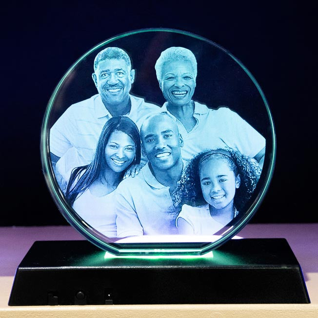 Bloc cube en verre gravé 3D représentant un couple souriant, posé en biais sur un socle rond rotatif LED bleue, idéal comme cadeau personnalisé lumineux.