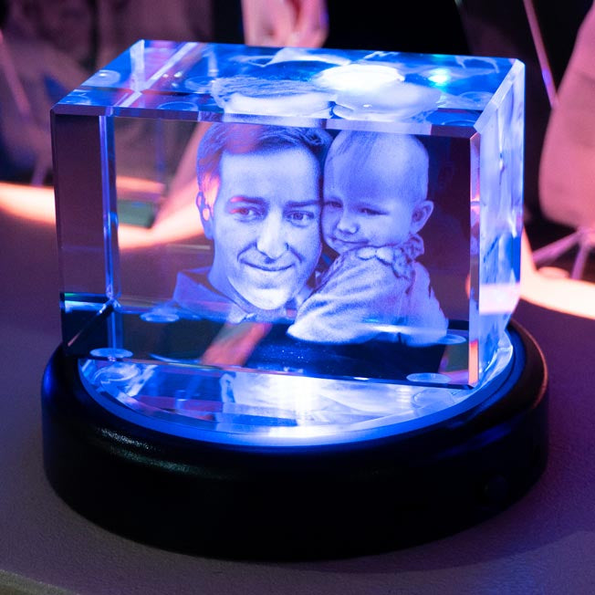 Un père tient son bébé dans un bloc en verre allongé, gravé en 3D et posé sur un socle lumineux. Ce bloc personnalisé est idéal pour la fête des pères ou une naissance.