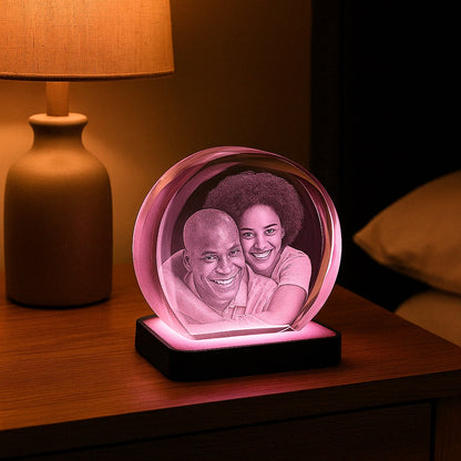 Bloc en verre rond gravé en 3D avec le portrait d’un couple, posé sur un socle lumineux diffusant une lumière rose, sur une table de chevet.