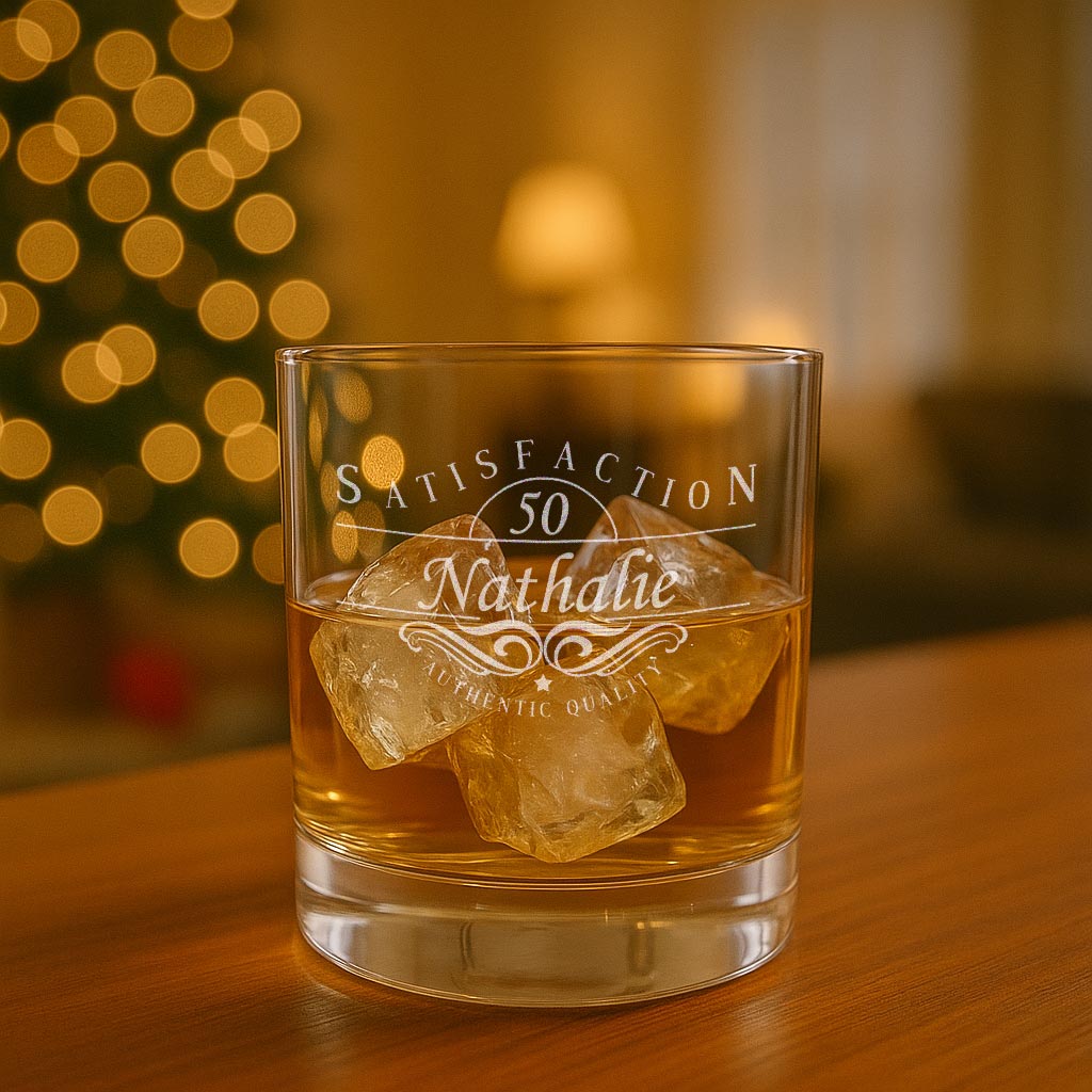Verre à whisky personnalisé avec l’inscription “Satisfaction 50 Nathalie” gravée au laser, posé sur une table dans une ambiance chaleureuse et festive.