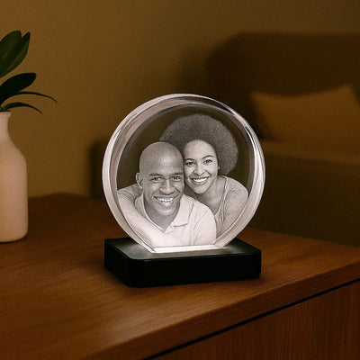 Bloc en verre rond gravé en 3D avec un portrait de couple, posé sur un socle lumineux blanc, sur une table de chevet en bois.