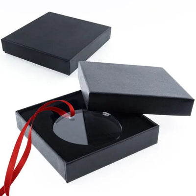 Boîte cadeau carrée noire ouverte, contenant un médaillon rond en verre avec un ruban rouge, sur fond blanc.