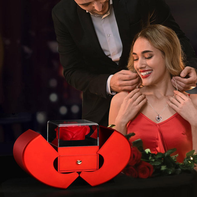 Mise en scène romantique : une femme reçoit un collier, devant un coffret cœur rouge avec rose éternelle. Idéal pour un cadeau d'amour.