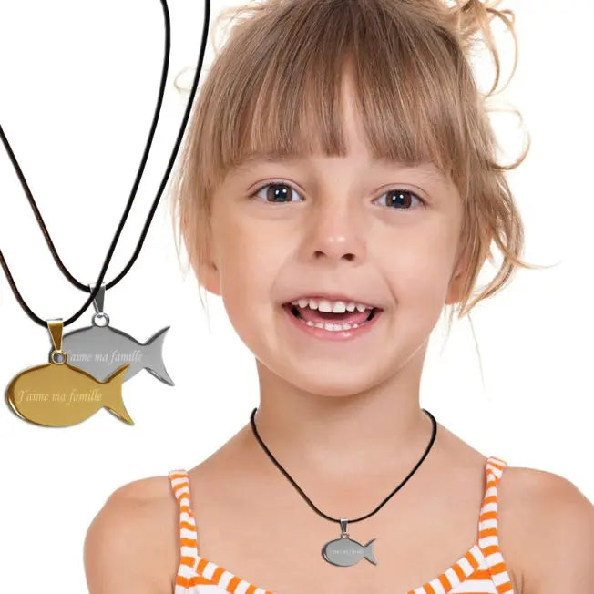 Petite fille souriante portant un pendentif poisson personnalisé en métal argenté, avec deux versions du bijou visibles en arrière-plan, doré et argenté.